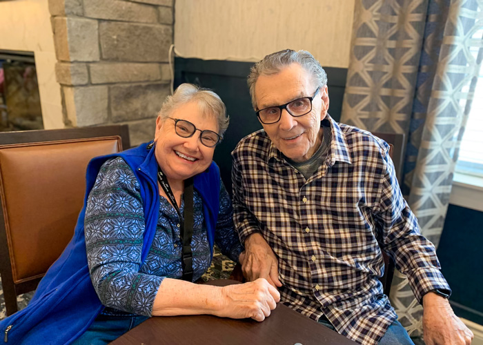 Two senior residents, both wearing glasses, smile while sitting together at a table in a senior living community, enjoying a moment of companionship.