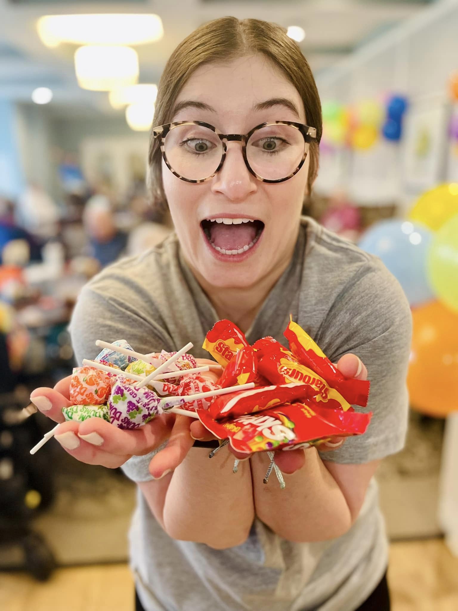 Excited team member with glasses holds an assortment of colorful candy, including Starburst and Dum Dums, during a fun event at a senior living community filled with balloons.