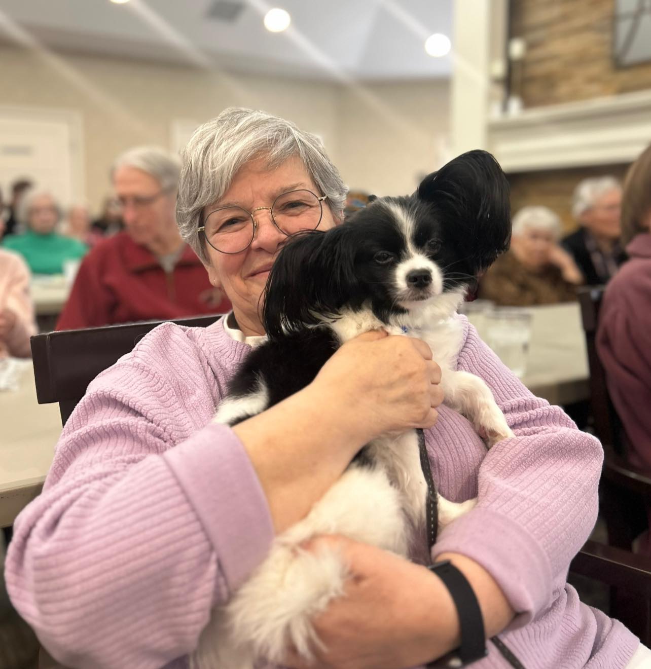 A senior resident wearing glasses and a lavender sweater smiles while holding a small black and white dog during an event at a senior living community.