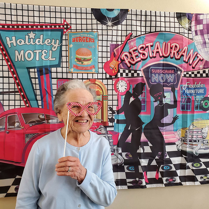 Smiling senior resident in a light blue sweater holds up a pair of heart-shaped glasses on a stick, posing in front of a colorful 1950s-themed diner backdrop with retro decor.