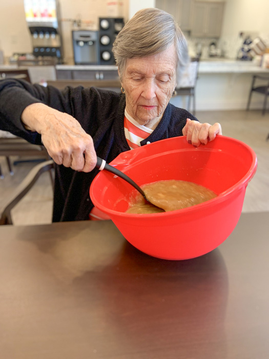 Senior resident in a senior living community carefully stirring a thick mixture in a large red bowl, fully focused on her task in a cozy kitchen setting.