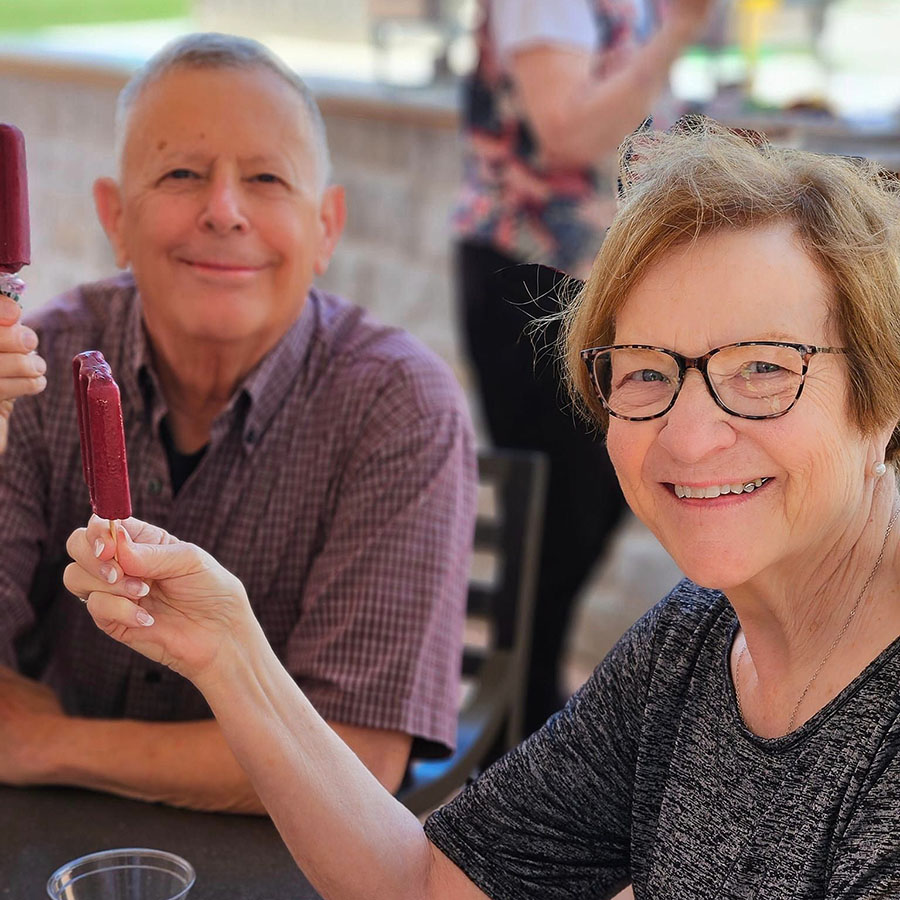 Smiling senior resident couple enjoys outdoor popsicles together on a sunny day, with the woman in glasses holding up a berry-flavored treat, celebrating a casual, happy moment.