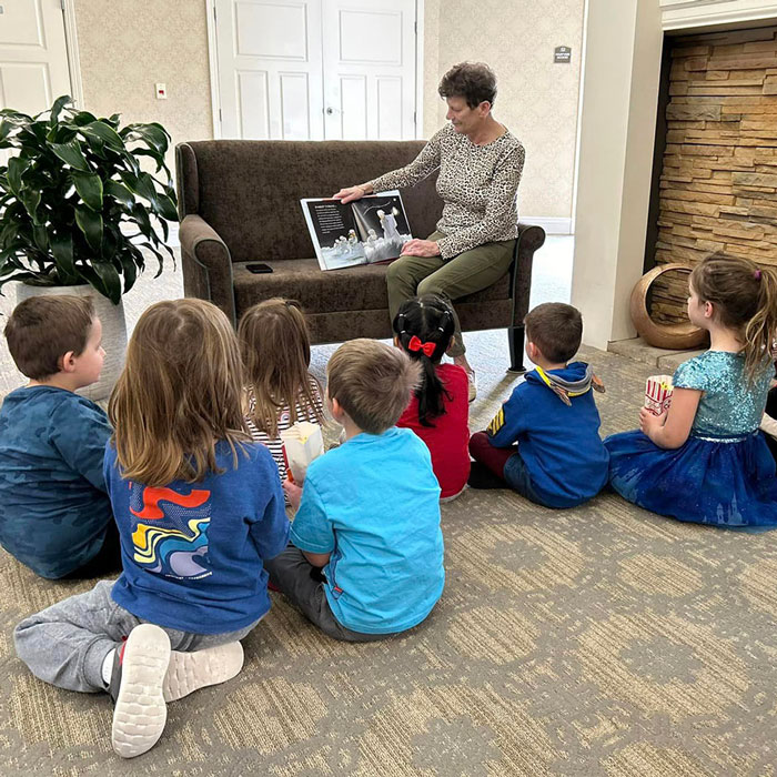 A senior resident reads a picture book to a group of attentive young children seated on the floor in front of a couch, during a storytime session in a cozy, well-lit room.