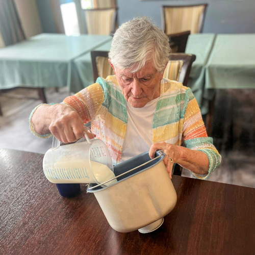 A senior resident with short gray hair pours liquid from a measuring cup into a mixing bowl, preparing to bake bread at a table in a senior residential dining area.