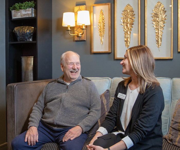 Smiling senior resident sitting on a couch and engaging in a joyful conversation with a staff member in a warmly lit room adorned with golden leaf wall art.