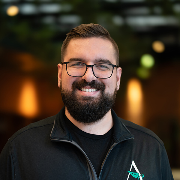 Johnathan Gallo, Executive Director at The Gardens at Arkanshire Senior Living, smiling warmly in a black jacket with an Arrow Senior Living logo, standing in a softly lit environment.