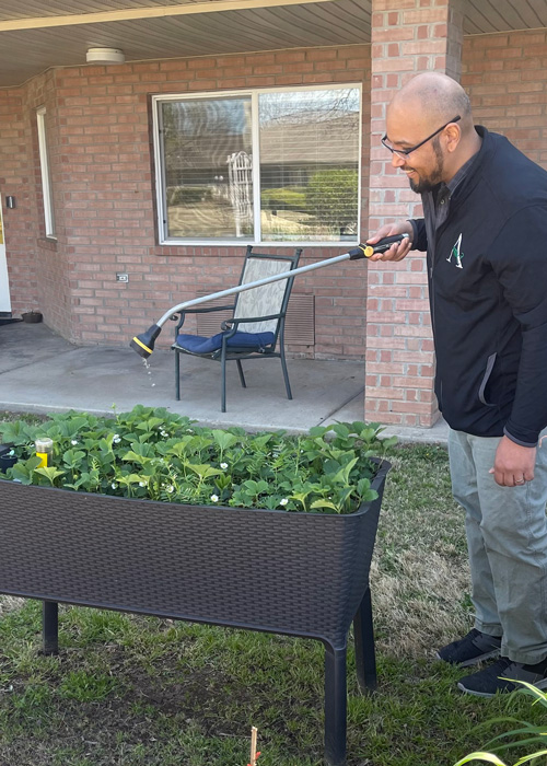 Smiling man in a black jacket uses a garden hose to water a raised bed of green plants outside a senior living community building.