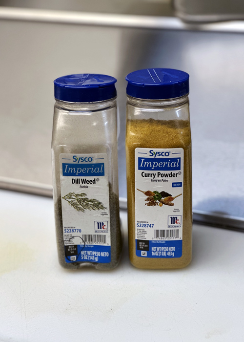 Sysco Imperial containers of dill weed and curry powder placed side by side on a kitchen prep counter, ready for use in seasoning and flavoring dishes.
