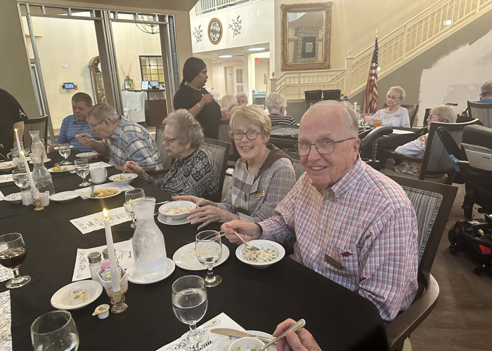 Group of senior residents enjoying a candlelit dinner with soup and drinks at a long banquet table inside a warmly lit senior living community.