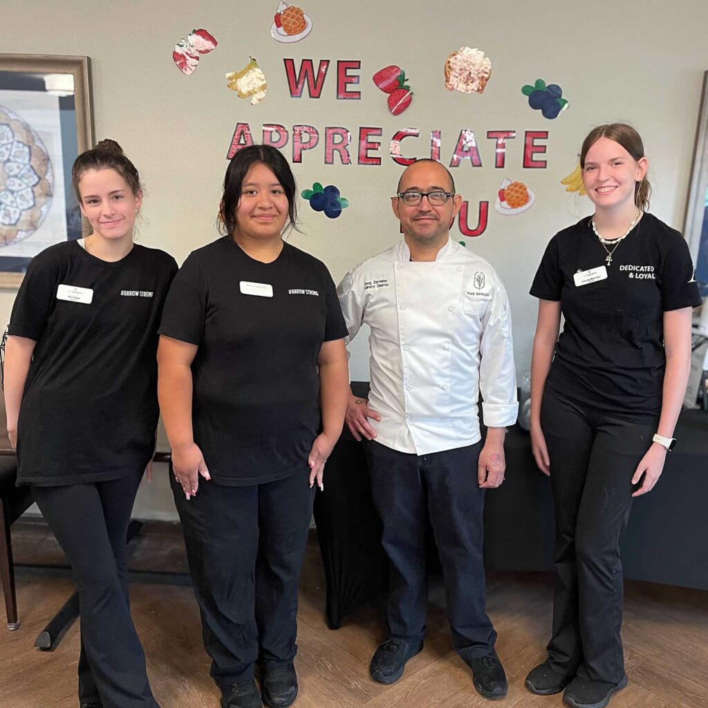 A chef in a white coat poses with three smiling team members in black uniforms beneath a colorful "We Appreciate You" sign at a senior living appreciation event.