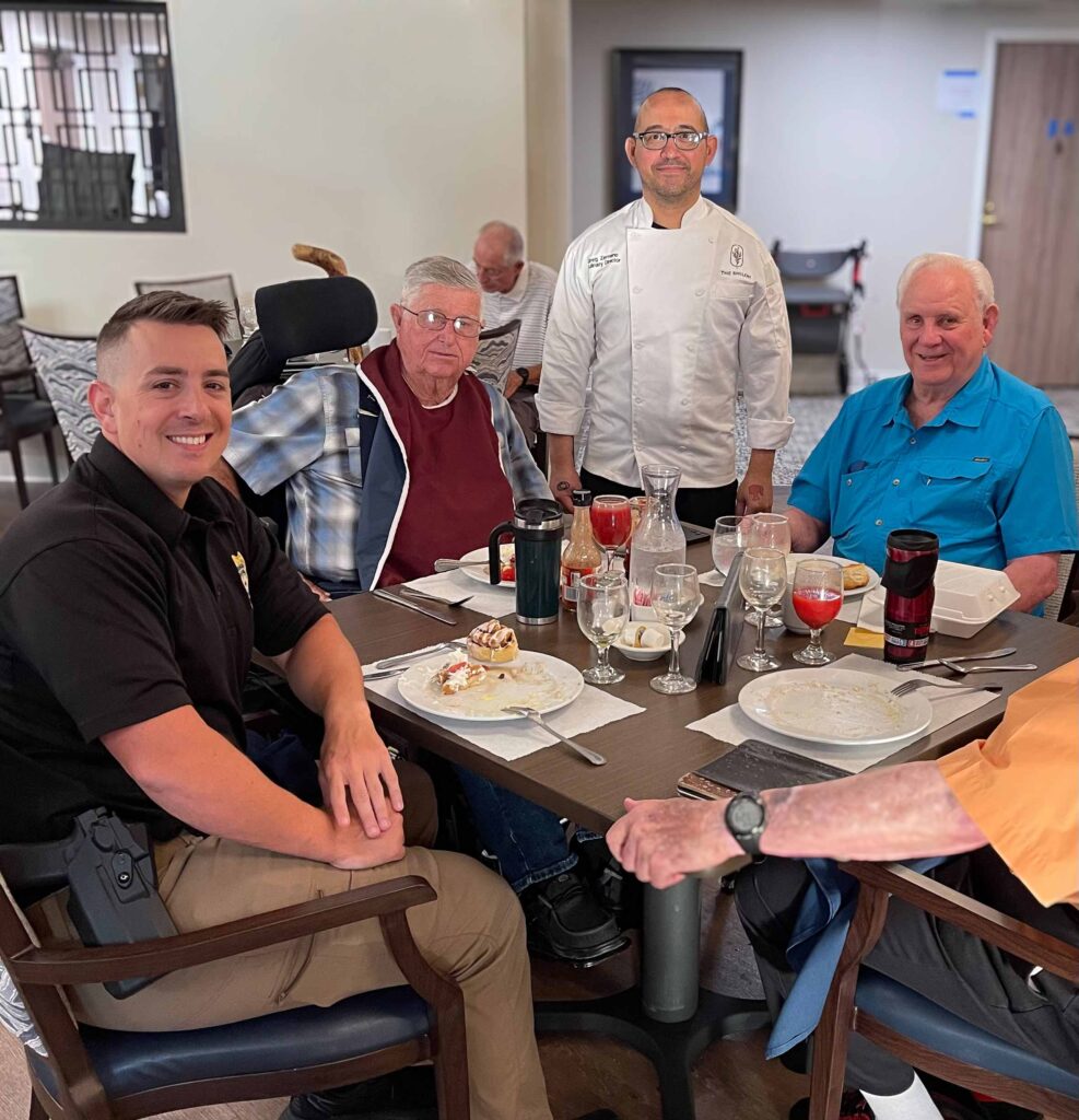 A chef in a white coat stands with residents and visitors at a dining table, enjoying dessert and drinks during a lively mealtime at a senior living community.