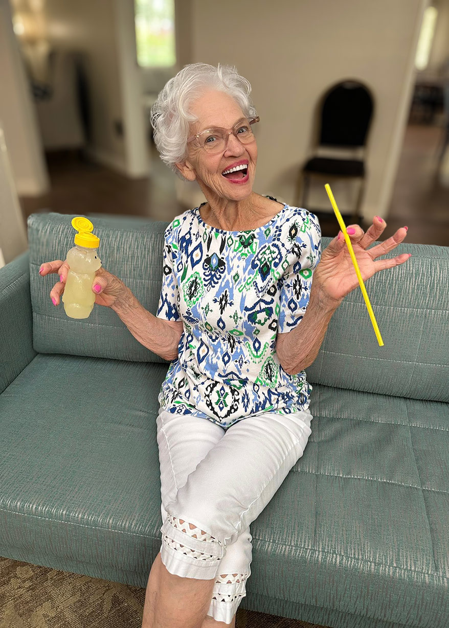 A senior woman smiling brightly sits on a sage couch, holding a honey bear with beverage inside, and yellow straw.