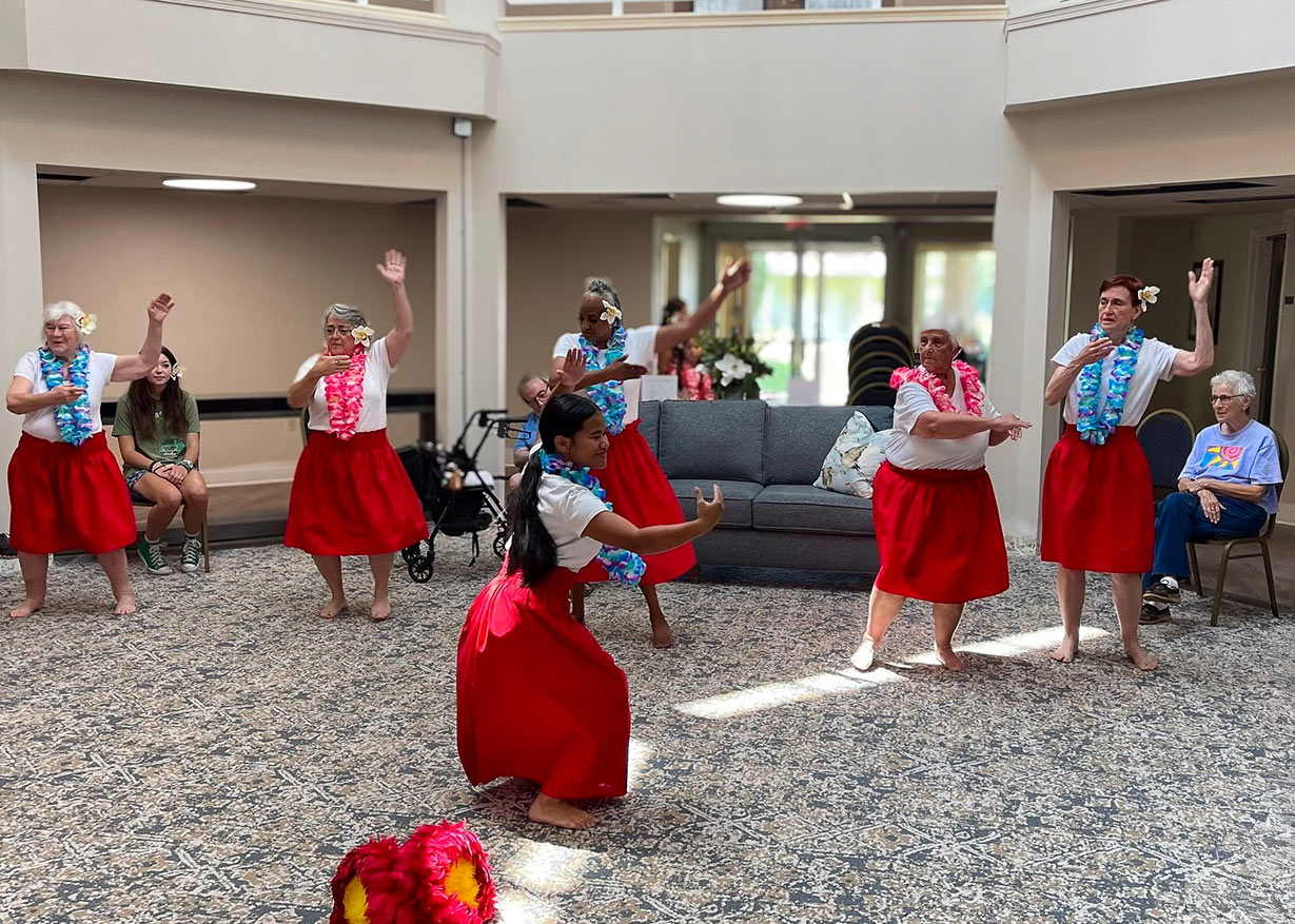Senior residents wearing red skirts and colorful leis perform a Hawaiian dance in a bright community living room, bringing joy and cultural celebration to the audience.