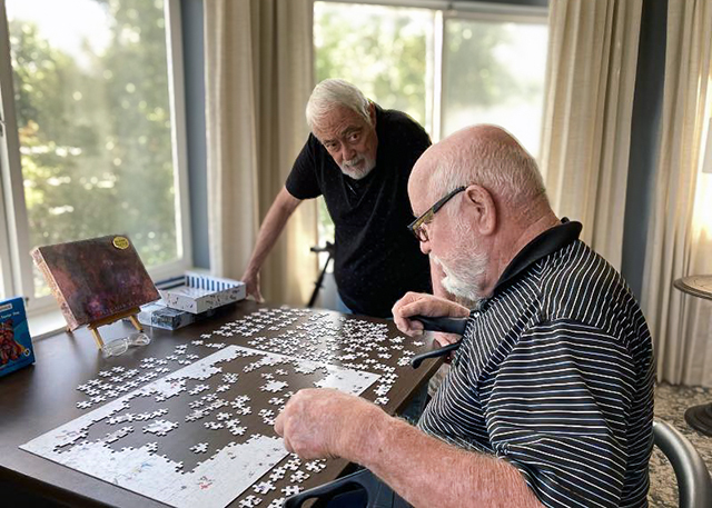 Two senior men consult one another while working on a large, intricate puzzle in a softly decorated room with large windows all around, bathed in natural light.