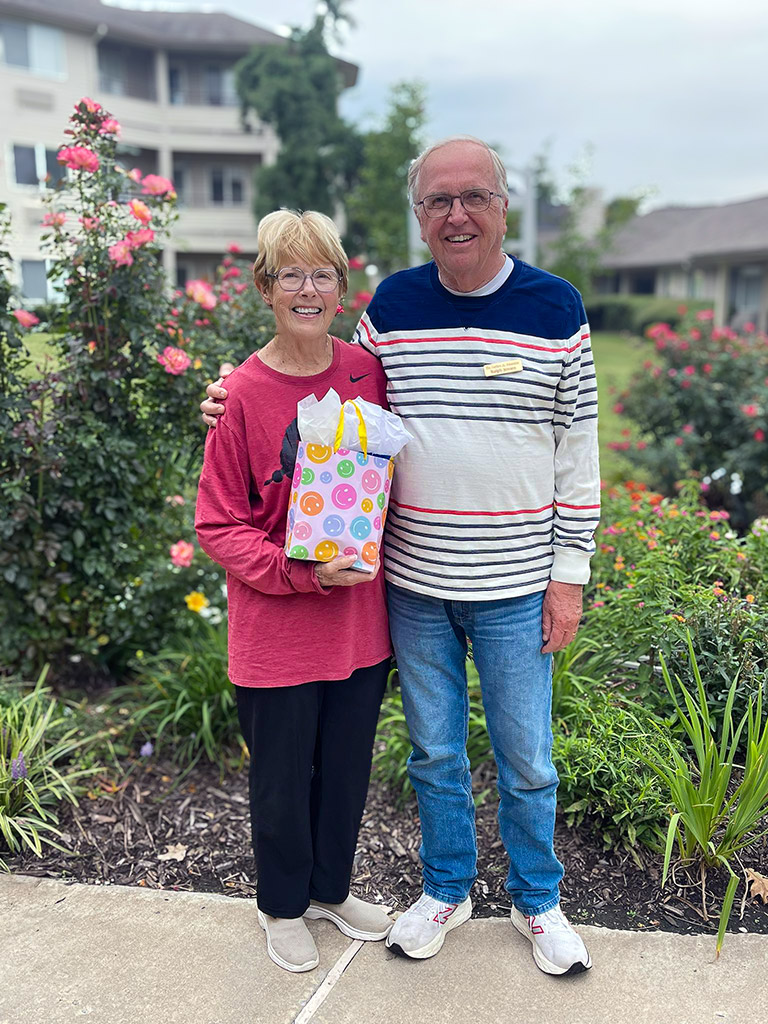 Senior man and woman stand together with a gift bag on a summer day on a foot path surrounded by roses, assorted flowers and ornamental grasses.