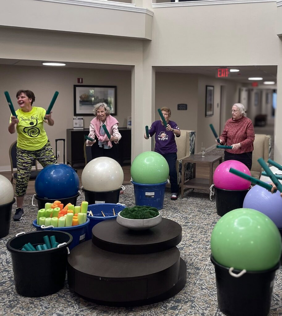 Residents seated in a circle with large, colorful exercise balls and drumsticks, participating in a lively fitness session.