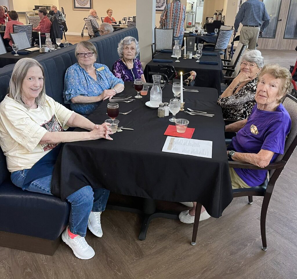 Several senior women share a tastefully set dining table, preparing to enjoy a chef-prepared meal at The Gardens at Arkanshire Senior Living.