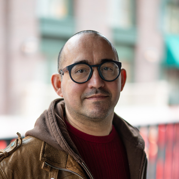 Gregorio “Greg” Zermeno, Culinary Director at The Gardens at Arkanshire Senior Living, posing in a professional headshot, wearing round glasses, a brown jacket, and a red sweater, with a softly blurred outdoor background.