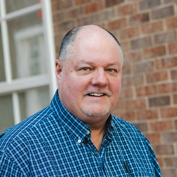 Rick Halbardier, Plant Operations Director at The Gardens Senior Living, smiling in a professional headshot, wearing a blue checkered button-down shirt, with a brick wall and window background.