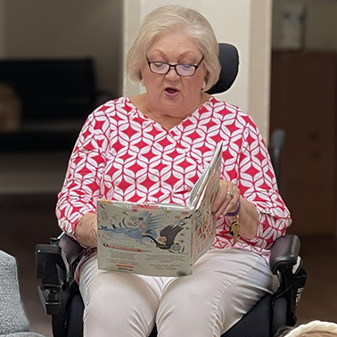 A senior woman wearing a red and white blouse reads a book to a group of children.
