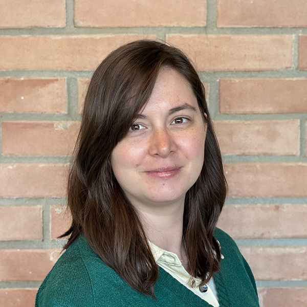 Aili Mendenhall, Senior Living Coordinator at The Gardens Senior Living, looking at the camera in a professional headshot with shoulder-length brown hair, a green sweater, and a brick wall background.